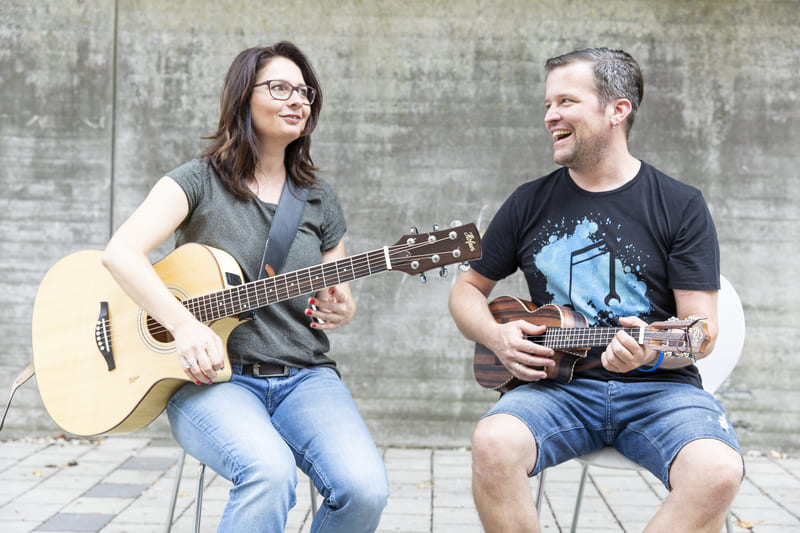 Die beiden Schulleiter Anita ite und Klaus Wüstner sitzen nebeneinander auf Stühlen und blicken sich an. Anita hält eine Gitarre in den Händen. Klaus hält eine Ukulele in den Händen.